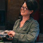 Frau mit grüner Bluse, dunklen Haaren und Brille lacht.