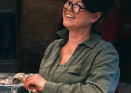 Frau mit grüner Bluse, dunklen Haaren und Brille lacht.