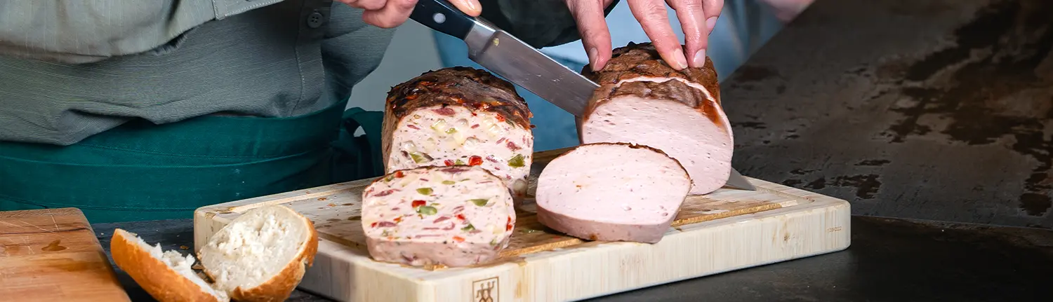 Ein aufgeschnittener Pizzaleberkäse und ein aufgeschnittener Leberkäse auf einem Holzbrett. Daneben ein aufgeschnittenes Brötchen.