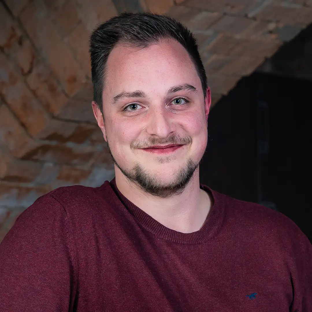 Andre Mayer aus dem Qualitätsmanagement der Metzgerei Lindörfer steht lächelnd vor einer rustikalen Ziegelwand. Er trägt ein bordeauxfarbenes Sweatshirt und blickt freundlich in die Kamera.