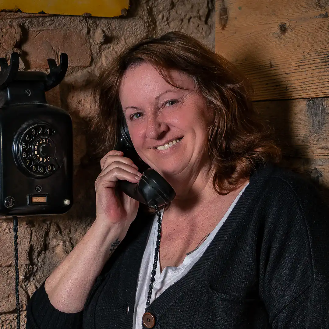 Bettina Marx vom Vertriebsinnendienst der Metzgerei Lindörfer lächelt freundlich, während sie einen schwarzen Retro-Telefonhörer ans Ohr hält. Sie steht vor einer Wand aus Holz und Naturstein.