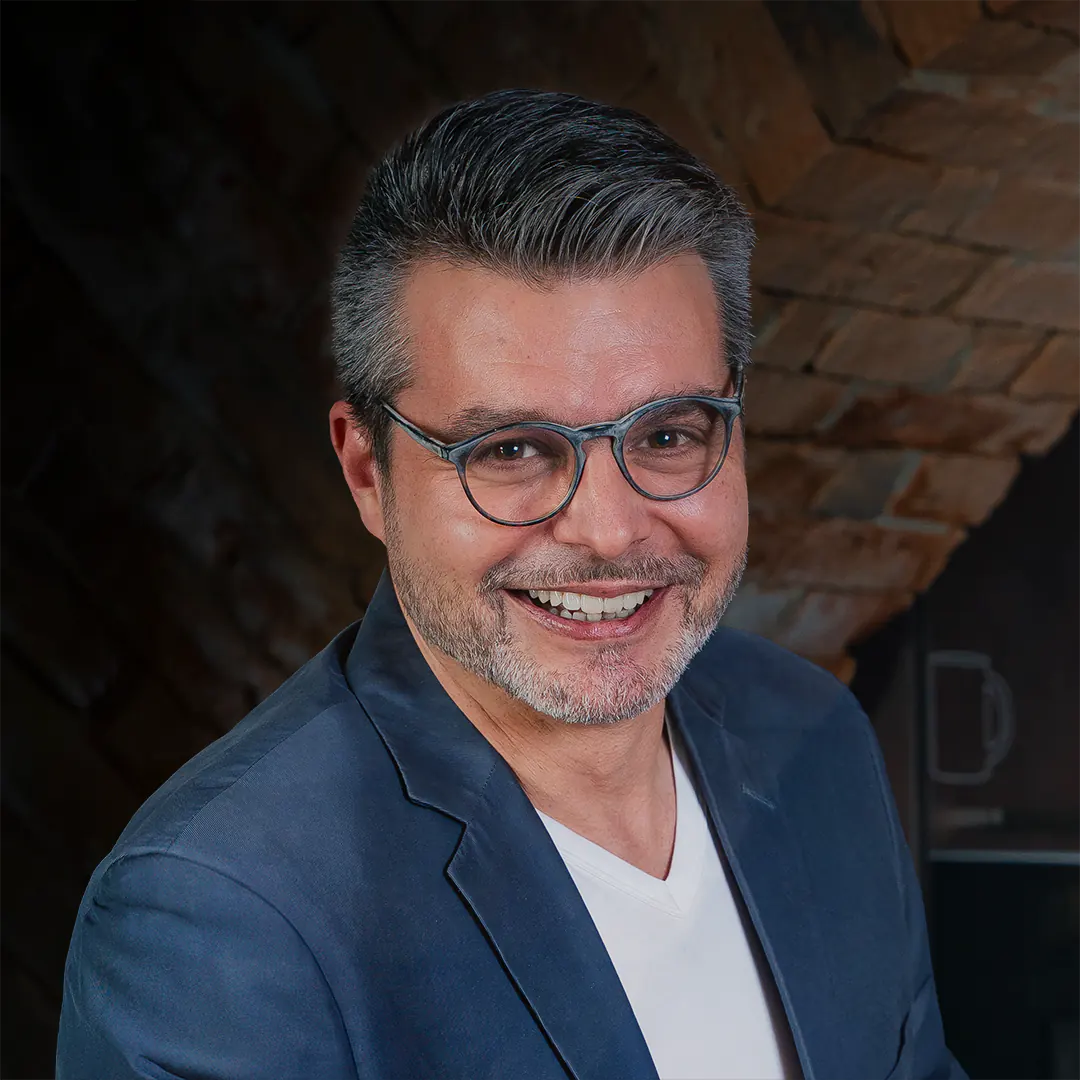 Christian Kohlschütter, Leiter der Verwaltung bei der Metzgerei Lindörfer, trägt eine Brille und ein dunkelblaues Sakko. Er lächelt freundlich in die Kamera vor einem gemauerten Hintergrund.