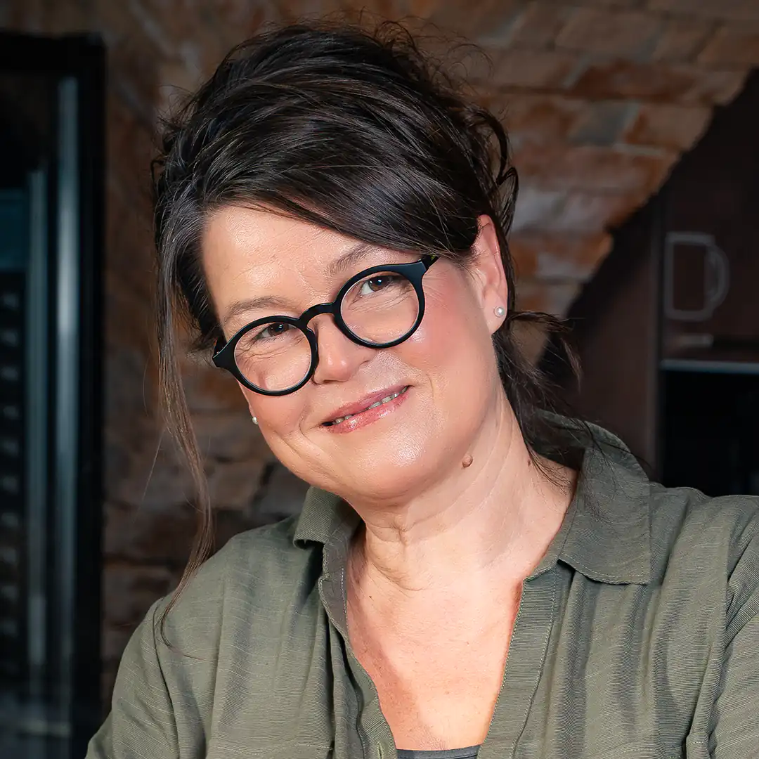 Jacqueline Kohlschütter, eine Frau mit dunklen, hochgesteckten Haaren und rundem Brillengestell, lächelt freundlich in die Kamera. Sie trägt eine olivgrüne Bluse und steht vor einer rustikalen Backsteinwand. Sie ist Geschäftsführerin der Metzgerei Lindörfer.