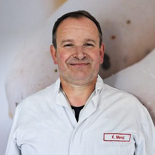 Karl Mend, Betriebsleiter der Metzgerei Lindörfer, trägt einen weißen Arbeitskittel mit Namensschild und steht vor einem Hintergrund mit Wurstmotiven. Er blickt freundlich in die Kamera.
