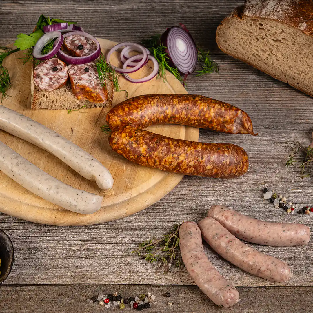 Auf einem runden Holzbrett liegen verschiedene rohe und gebratene Bratwürste: zwei helle, zwei gebräunte mit grober Struktur und vier kleine ungebratene Würste. Daneben eine Brotscheibe mit aufgeschnittener Wurst, rote Zwiebelringe, frische Kräuter, Pfefferkörner und ein Laib Bauernbrot auf einem hölzernen Untergrund.
