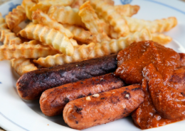 Drei gebratene vegane Bratwürste mit roter Soße und Pommes Frites auf weißem Teller.