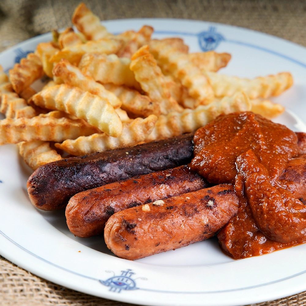 Drei gebratene vegane Bratwürste mit roter Soße und Pommes Frites auf weißem Teller.