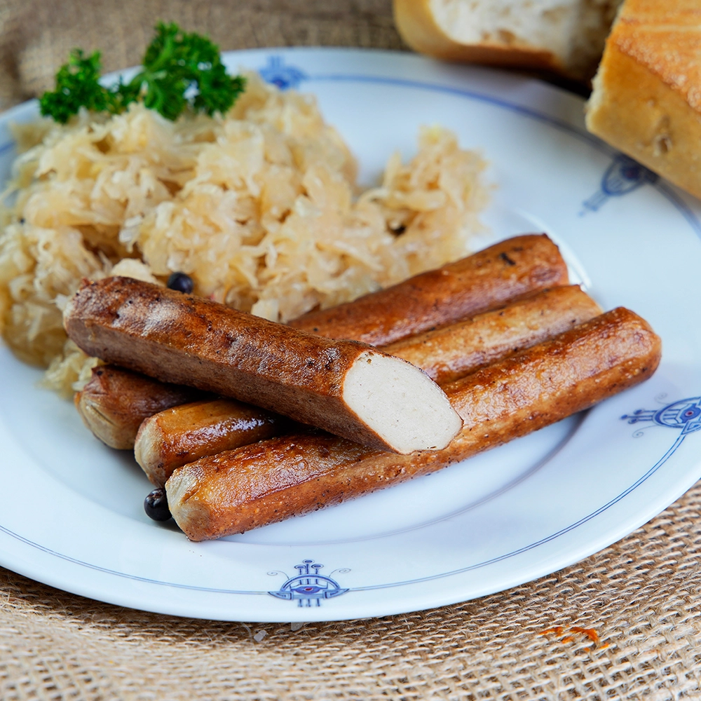 Eine angeschnittene Bratwurst auf drei weiteren gebratenen Bratwürsten neben Sauerkraut auf weißem Teller.