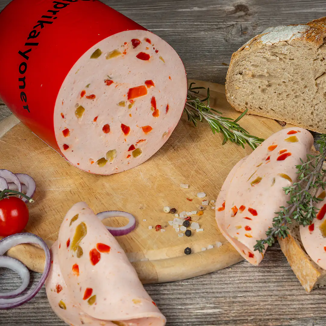 Eine angeschnittene große Paprikalyoner mit rotem Kunststoffmantel liegt auf einem runden Holzbrett. Die Wurst enthält rote und grüne Paprikastückchen. Daneben mehrere aufgerollte Scheiben, eine Scheibe Brot, rote Zwiebelringe, eine Cocktailtomate, Rosmarin, Thymian sowie grobes Salz und Pfefferkörner auf einem Holztisch.