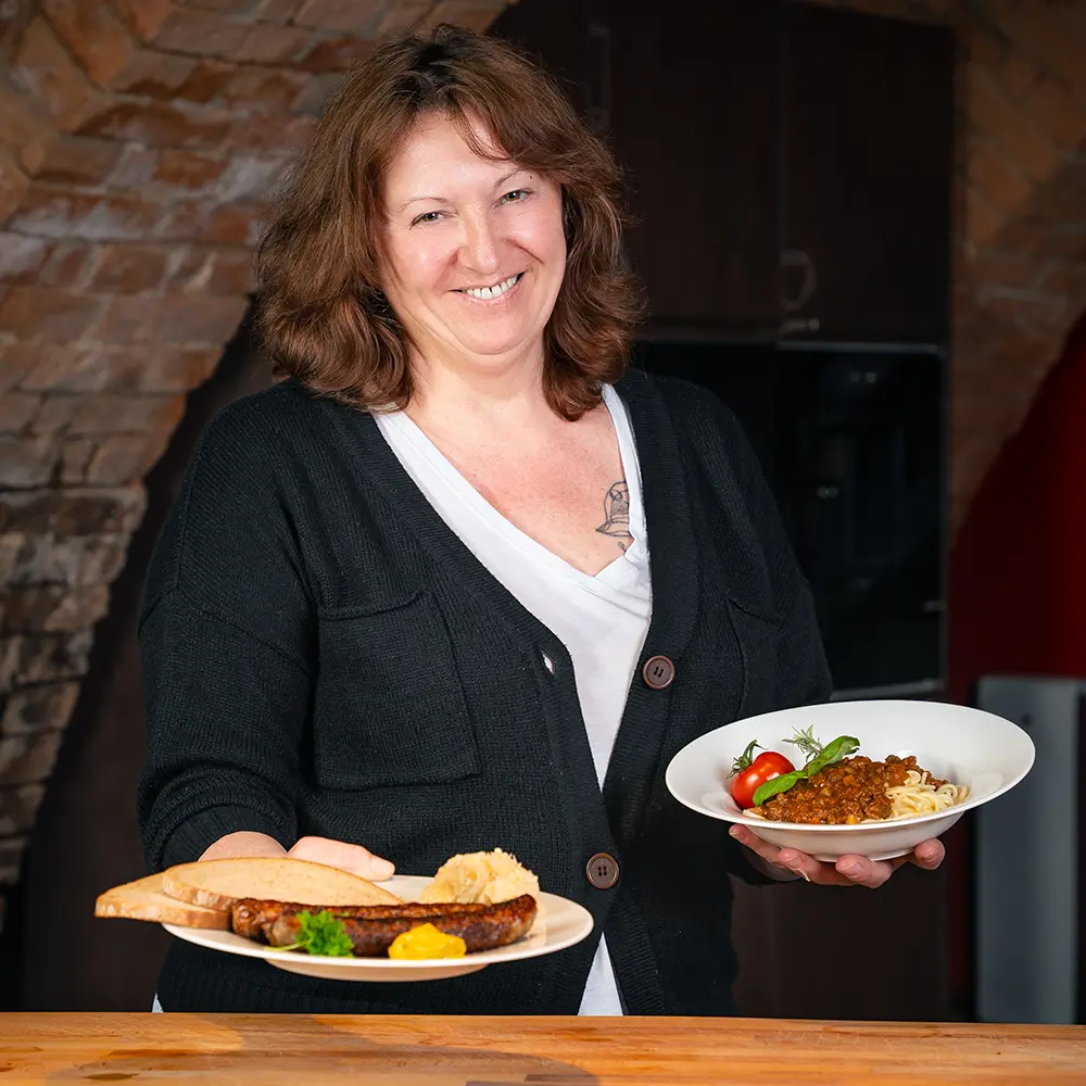Eine lächelnde Frau mit braunen Haaren, weißer Bluse und schwarzer Strickjacke hält zwei Teller it deftigen Speisen.