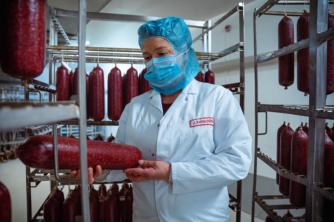 Eine Frau in weißem Kittel mit Namensschild ‚J. Kohlschütter‘ steht in einem Reiferaum für Salami. Sie trägt eine blaue Haube, eine medizinische Maske sowie einen Mundschutz und hält eine rote Rohwurst in der Hand. Im Hintergrund hängen zahlreiche Salamis in Reihen an Metallgestellen