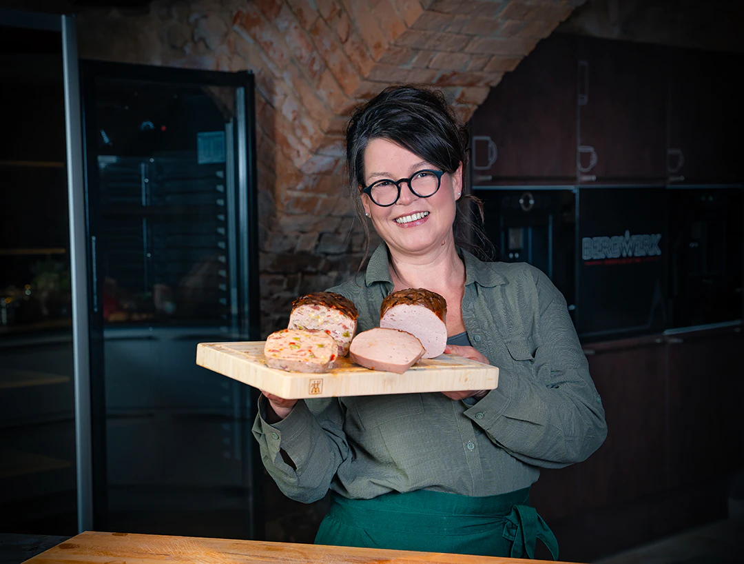Jacqueline Kohlschütter steht lächelnd in einem Raum mit Backstein-Gewölbedecke und hält ein Holzbrett mit drei angeschnittenen Fleischkäsevarianten in die Kamera. Sie trägt eine Brille, eine grüne Bluse und eine grüne Schürze. Im Hintergrund sind dunkle Küchenschränke zu sehen