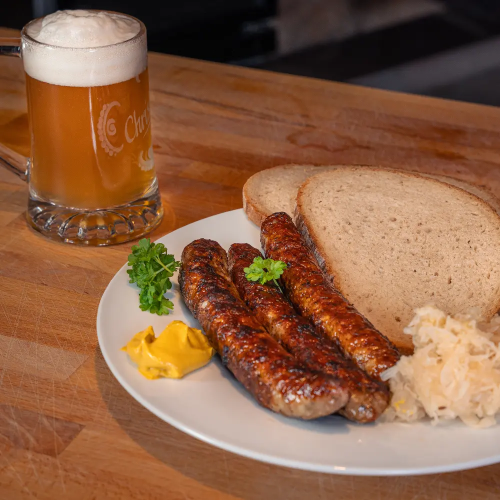 Drei gebratene Bratwürste mit zwei Scheiben Brot und Sauerkraut auf einem weißen Teller neben einem Krug mit Bier auf heller Holzplatte.