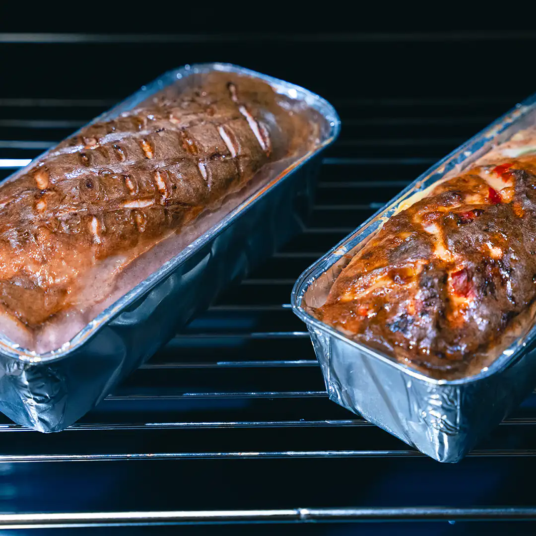 Zwei Leberkäse-Laibe in silberfarbenen Einwegbackformen stehen nebeneinander auf einem Ofengitter. Die Oberfläche ist goldbraun gebacken, einer der Laibe zeigt eine aufgebrochene Kruste mit leicht rötlicher Füllung.