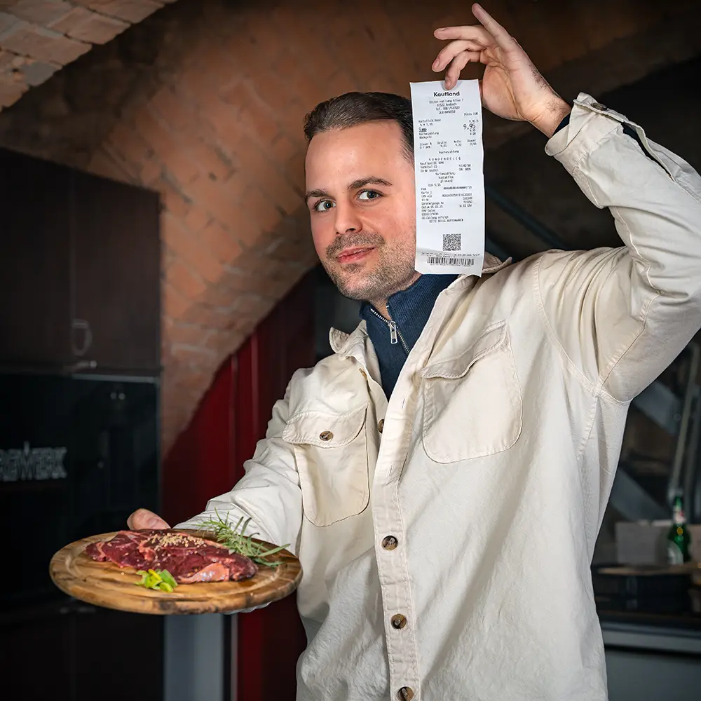 Ein Mann mit kurzen braunen Haaren und herausforderndem Blick hält mit der linken Hand einen Kassenbon nach oben. Ind er rechten Hand hält er eine runde Holzplatte mit rohem Fleisch.
