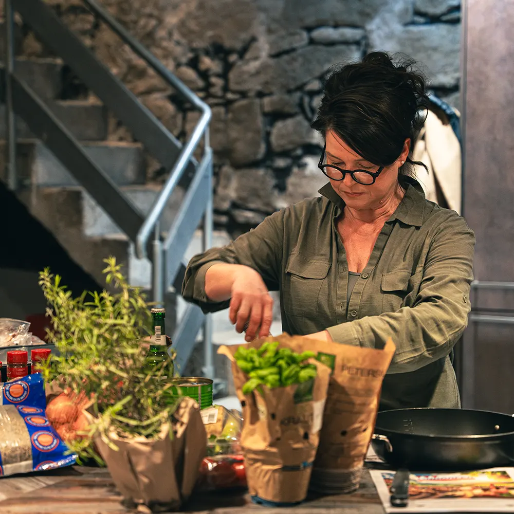Frau mit dunklen Haaren und grüner Bluse steht hinter verschiedenen Gewürzpflanzen und kocht.