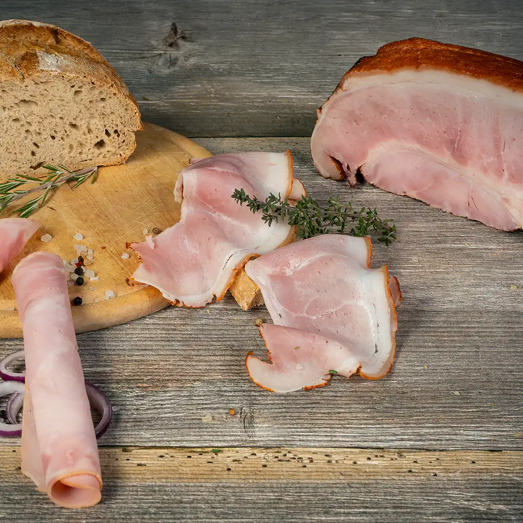 Mehrere Scheiben heller, rosa gekochter Schinken liegen auf einem rustikalen Holztisch, teils auf Brot, garniert mit Thymian und Rosmarin. Ein größerer ungeschnittener Schinkenblock sowie ein Laib Bauernbrot sind ebenfalls zu sehen. Daneben grobes Salz, Pfefferkörner und rote Zwiebelringe.