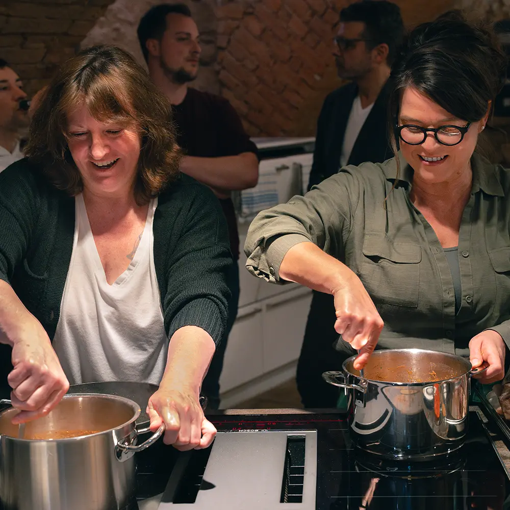 Zwei Frauen stehend lachend hinter Kochtöpfen und rühren darin mit einem Kochlöffel. Im Hintergrund unterhalten sich zwei Männer.