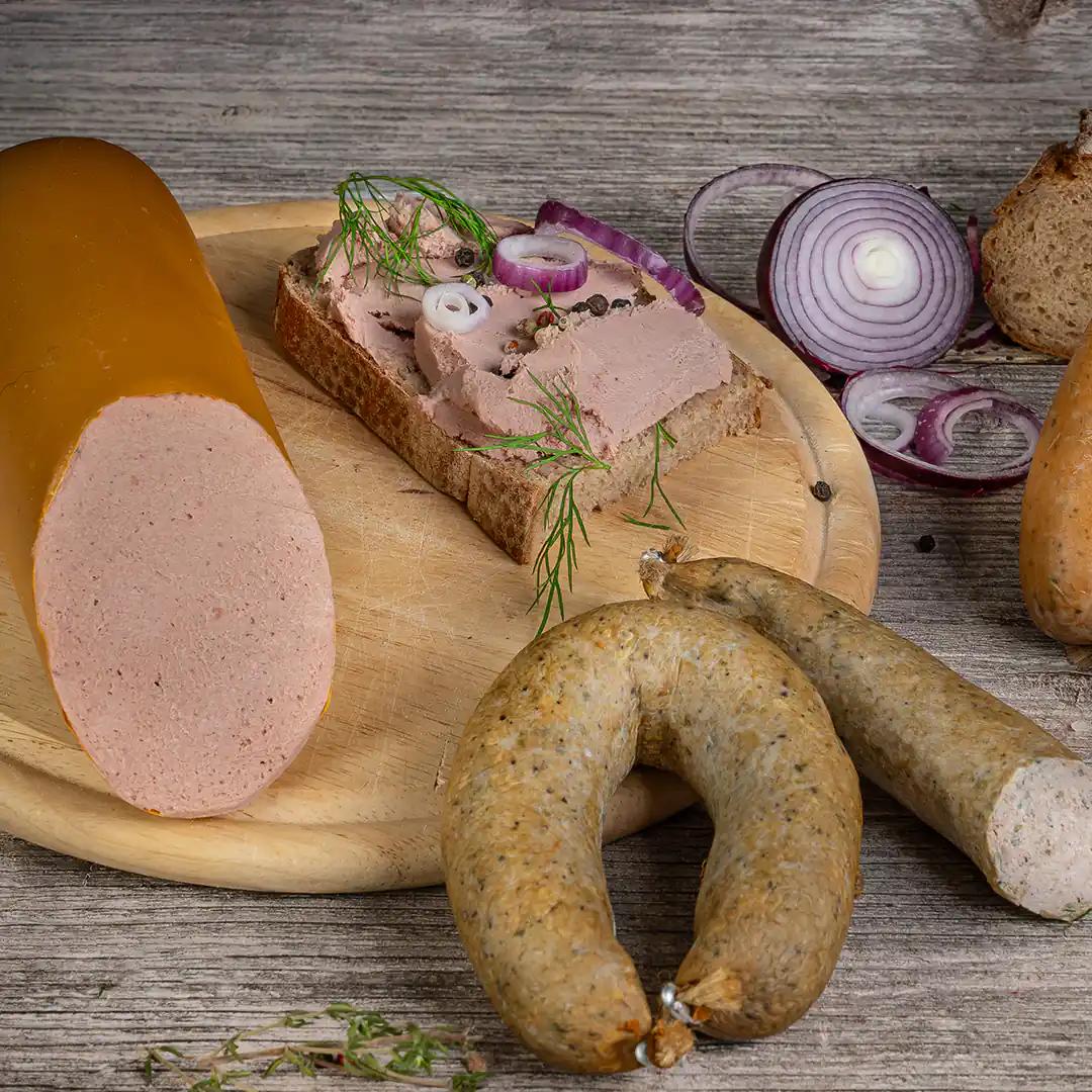 Auf einem Holzbrett liegt eine angeschnittene Streichwurst mit glatter, rosa Oberfläche und gelblicher Hülle. Daneben eine Brotscheibe mit aufgestrichener Leberwurst, dekoriert mit Zwiebelringen, Dill und Pfefferkörnern. Zwei grünlich-braune, grobe Würste mit Naturdarm liegen ebenfalls auf dem Brett und dem Tisch, umgeben von roten Zwiebelringen, Kräutern und einem Laib Bauernbrot.