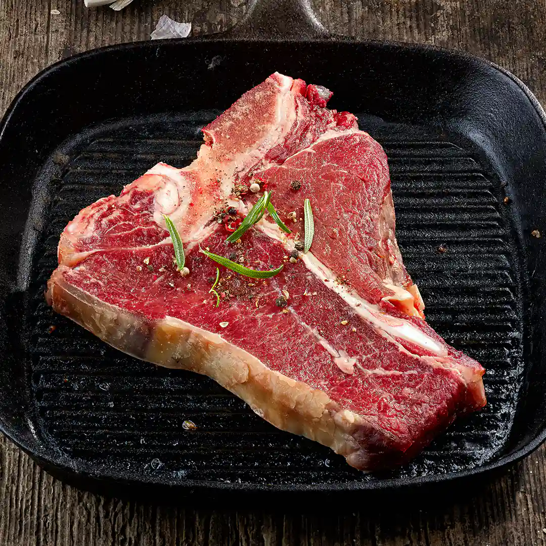 Rohes T-Bone-Steak gewürzt mit Pfeffer und Rosmarin, liegt in einer gusseisernen Grillpfanne auf rustikalem Holztisch.