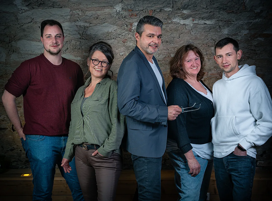Fünf Personen stehen nebeneinander vor einer rustikalen Steinwand und blicken in die Kamera. Von links nach rechts: ein Mann in dunkelrotem Shirt und Jeans (André Mayer, Qualitätsmanagement), eine Frau mit Brille und grünem Hemd (Jacqueline Kohlschütter, Geschäftsführung), ein Mann im grauen Sakko mit Brille in der Hand (Christian Kohlschütter, Leiter Verwaltung), eine Frau mit mittellangem Haar und schwarzem Cardigan (Bettina Hacker, Vertriebsinnendienst) sowie ein Mann in weißem Kapuzenpullover (nicht namentlich genannt). Alle lächeln freundlich.“
