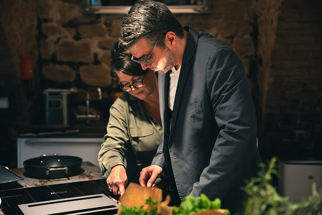 Mann mit kurzen Haaren und Dreitagebart und Brille und Frau mit grüner Bluse und Brillen stehen seitlich fotografiert zusammen am Herd und kochen.