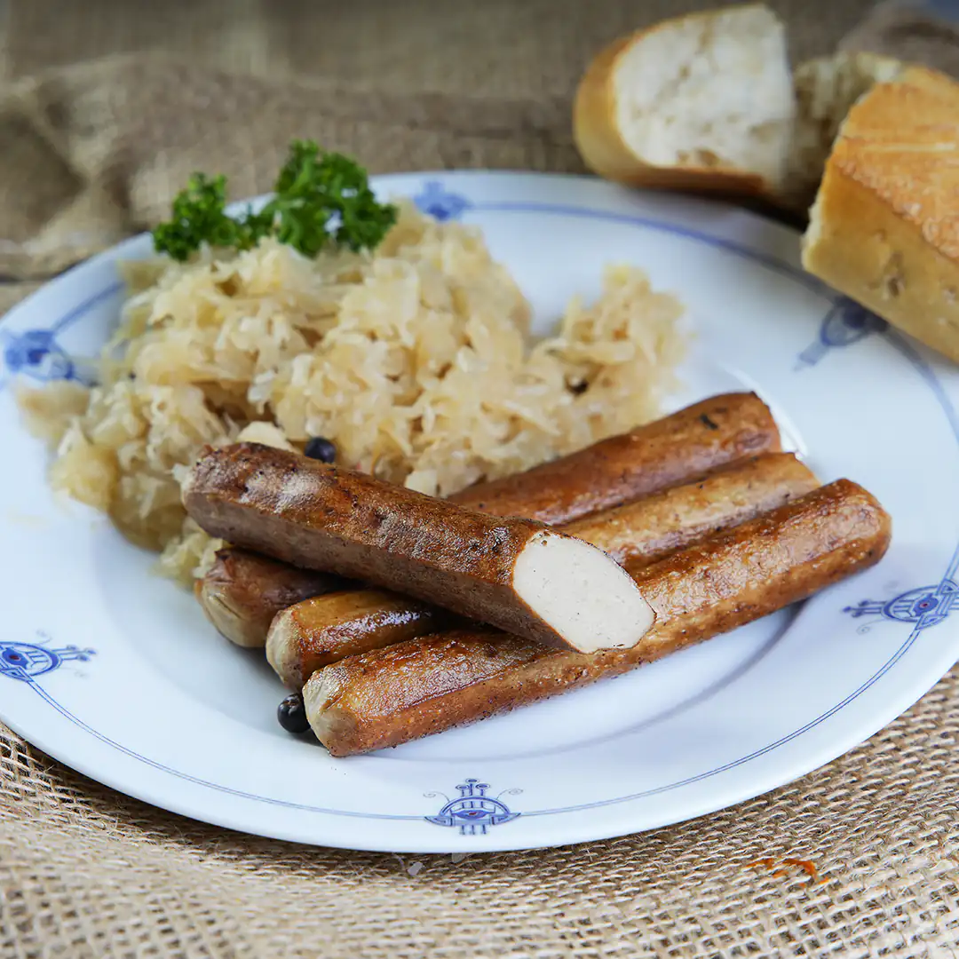 Auf einem weißen Porzellanteller mit blauem Muster liegen mehrere gebratene vegane Würstchen, eine davon ist angeschnitten. Dahinter befindet sich eine Portion Sauerkraut, garniert mit Petersilie. Im Hintergrund liegen zwei aufgebrochene Stücke Baguette auf einer Leinentischdecke