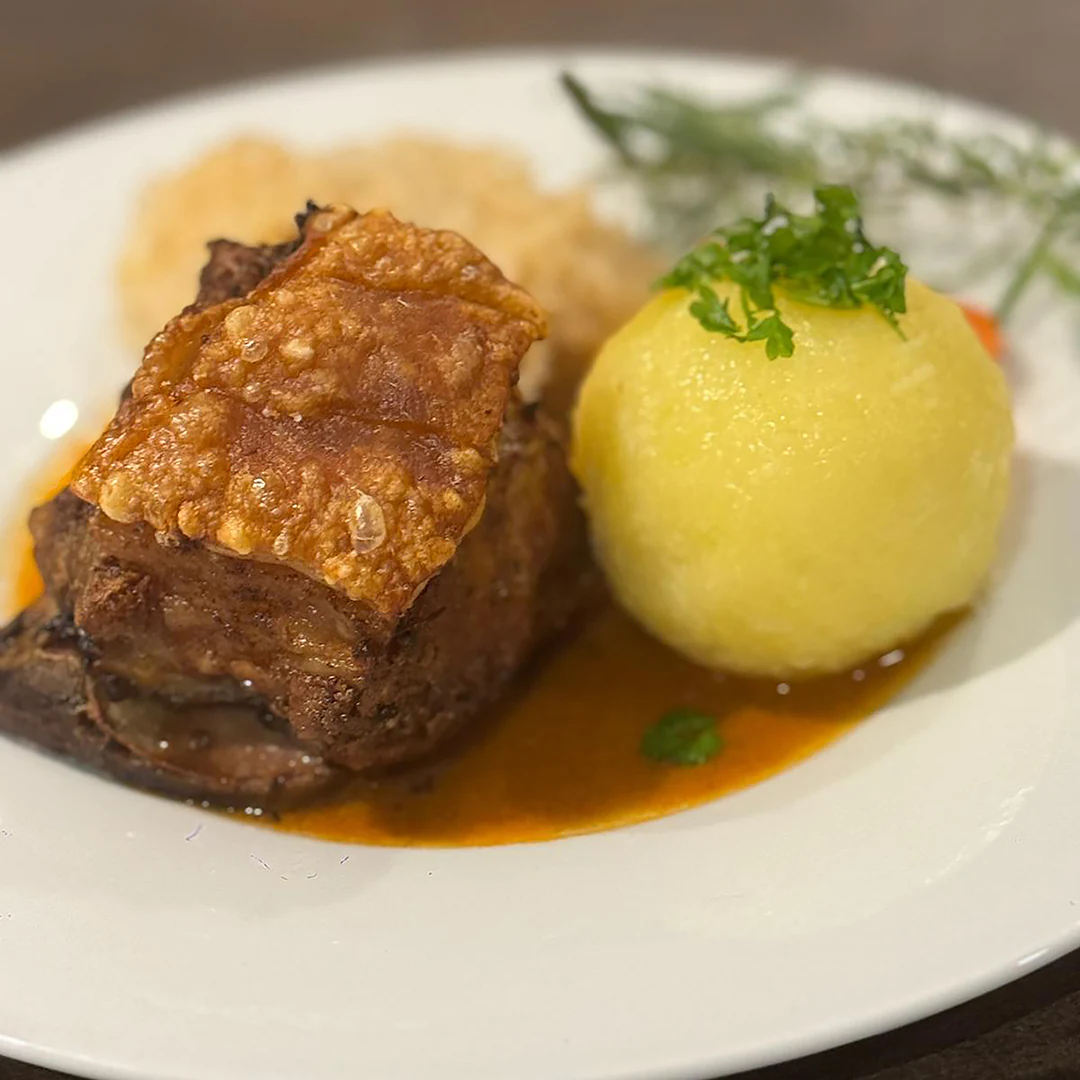 Auf einem weißen Teller liegt ein Stück Schweinebraten mit knuspriger Kruste in dunkler Soße, daneben ein runder Kartoffelknödel mit etwas Petersilie sowie eine Portion heller Krautsalat im Hintergrund. Der Teller steht auf einem dunklen Untergrund